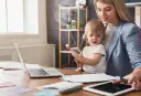 Pracująca mama z dzieckiem na kolanach, laptop i tablet na biurku. Czy na macierzyńskim można pracować? Tak, to możliwe!
