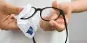 Dłonie czyszczą okulary okularowe białą ściereczką z logo Zeiss.