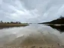 Spokojne jezioro Wójtowskie pod pochmurnym niebem. Woda odbija chmury i zarośla trzcin.