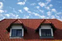 Czerwony dach z dachówki ceramicznej z dwoma lukarnami na tle błękitnego nieba z chmurami.