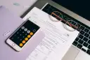 Telefon z kalkulatorem, okulary i dokumenty podatkowe na laptopie.