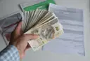 Dłoń trzyma plik banknotów 200 zł. W tle dokumenty, być może dotyczące tego, ile godzin to 0.5 etatu.