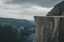 Człowiek w żółtej kurtce stoi na krawędzi skalnego klifu w Norwegii, podziwiając majestatyczny fiord i góry.