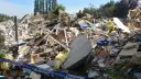 Ogromna kupa gruzów po zawaleniu się budynku. Widać izolację, drewno, beton i niebieską torbę. Wstęga policyjna zabezpiecza teren.