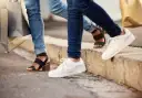 Dwie osoby w jeansach i modnych butach, z torbami na zakupy w rękach, idą chodnikiem. Jedna ma czarne sandały na obcasie, druga białe sneakersy.