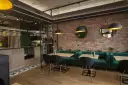 Nowoczesne wnętrze restauracji z ceglaną ścianą, zielonymi kanapami i drewnianymi stołami.