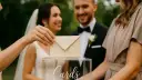 Dłoń wkłada kopertę do przezroczystej skrzynki z napisem "Cards". Młoda para w tle uśmiecha się, a goście wrzucają koperty, być może z myślą, ile na prezent ślubny.