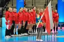 Drużyna piłki ręcznej kobiet Polski w czerwonych strojach stoi w rzędzie, trzymając polską flagę.