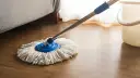 Mop z białymi sznurkami na drewnianej podłodze, obok wiaderka. Idealny do pytania: czym myć panele.