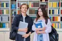 Dwóch studentów z książkami i laptopem na tle regałów z książkami.