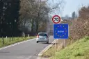 Znak graniczny Belgii, gdzie potrzebny paszport, na tle drogi i jadącego samochodu.