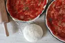 Dwa surowe ciasta na pizzę z sosem pomidorowym i ziołami, wałek i kulka ciasta.
