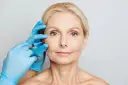 Zabieg z użyciem botoksu, czy botox jest bezpieczny? Dłoń w niebieskiej rękawiczce wykonuje zastrzyk w okolicę oka kobiety.