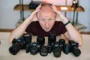 Mężczyzna z rękami na głowie otoczony aparatami różnych marek: Sony, Canon, Fujifilm.