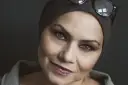 Sylwia Janowicz-Dobrowolska w szarej opasce i okularach na głowie, z wyrazistym makijażem oczu i błyszczącymi ustami.