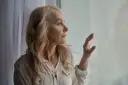 Starsza kobieta z blond włosami patrzy przez okno, zamyślona.