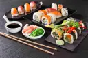 Różnorodne sushi, sos sojowy, marynowany imbir, sałatka wakame i pałeczki na ciemnym tle.