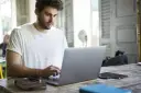 Mężczyzna w białej koszulce pisze na laptopie, przygotowując wniosek o urlop na poszukiwanie pracy.