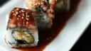 Rolka sushi z wężem morskim, polana słodkim sosem unagi i posypana sezamem, leży na talerzu.