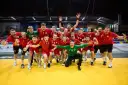Drużyna piłkarzy ręcznych w czerwonych strojach świętuje zwycięstwo. Wśród nich Krzysztof Zimny, bramkarz w zielonym stroju, klęczy z wyciągniętymi rękami.