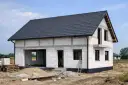 Budowa domu ze styropianu w stanie surowym. Białe bloczki, szary dach, okna i drzwi czekają na wykończenie.