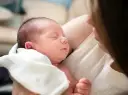 Śpiący noworodek wtulony w ramiona mamy. Delikatne światło podkreśla jego rysy.