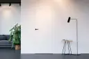 Białe drzwi ukryte w ścianie z czarną klamką, obok stojąca lampa i stolik.