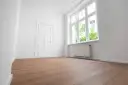 Puste, jasne pomieszczenie z białymi ścianami, drzwiami i oknem. Drewniana podłoga i grzejnik sugerują, że to gotowe do zamieszkania mieszkanie socjalne.