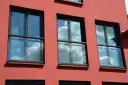 Trzy nowoczesne okna z czarnymi ramami i żaluzjami, odbijające błękitne niebo z chmurami. Wyglądają jak małe balkony francuskie.