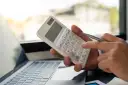 Dłoń z długopisem naciska klawisz kalkulatora, pomagając obliczyć zużycie prądu obok laptopa.