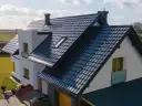 Nowoczesny dom z białymi ścianami i ciemnym, błyszczącym dachem dwuspadowym. Widać okna, komin i fragment ogrodu.
