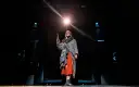 Dziewczynka z zapałkami na scenie, trzymająca płonącą zapałkę, w otoczeniu mroku i teatralnego światła.
