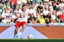 Jakub Kozłowski w akcji, w białej koszulce z orłem i czerwonych spodenkach, prowadzi piłkę na boisku.