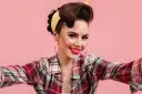 Uśmiechnięta kobieta w stylu pin-up, z burzą brązowych włosów spiętych w kok i żółtą bandaną, w kratę koszuli, robi sobie selfie.