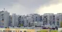 Rząd szarych bloków z wielkiej płyty, z balkonami i antenami, pod pochmurnym niebem.