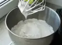 Ubita śmietana z mascarpone, puszysta i lśniąca, spływa z mieszadła miksera do metalowej misy.
