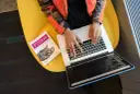 Osoba w pomarańczowej kurtce pisze na laptopie obok książki o Pythonie, siedząc na żółtym krześle.