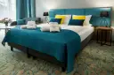 Przytulny pokój hotelowy z dużym łóżkiem, ozdobionym turkusową narzutą i żółto-niebieskimi poduszkami. Ręczniki z logo Stella Resort & Spa zachęcają do relaksu.