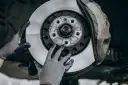 Mechanik w rękawiczkach wymienia zużyte tarcze hamulcowe. Widać nowy, lśniący element obok starego zacisku.