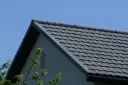 Szary dach z dachówki ceramicznej pod błękitnym niebem, z fragmentem ściany domu i gałęziami drzew.
