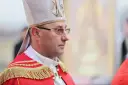 Biskup w stroju pontyfikalnym, z mitrą na głowie, w okularach. W tle widać fragment obrazu.
