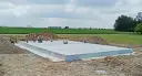 Gotowa płyta fundamentowa na budowie, otoczona piaskiem i zielenią.