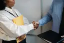 Uścisk dłoni po rozmowie o przedłużenie umowy o pracę. Kobieta trzyma żółtą teczkę, mężczyzna czarną.