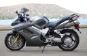 Szary motocykl Honda VFR 800 z widokiem na wodę i wzgórze w tle.