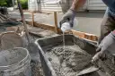 Rękawiczki i łopatka mieszają szybki beton w wózku. Obok leży worek z cementem i wiadro.