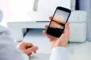 Dłoń trzyma smartfon z napisem "Sending to printer", pokazując jak drukować z telefonu. W tle biała drukarka.