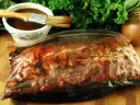 Karkówka w torebce, gotowa na grilla. W miseczce czeka aromatyczna marynata do karkówki na grilla, a obok pęczek zielonej sałaty i cebula.