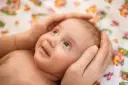 Delikatne dłonie otulają główkę dziecka, chroniąc jego miękkie ciemiączko. Maluch patrzy z zaciekawieniem w górę, uśmiechając się lekko.