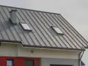 Nowoczesny dach z szarej blachy na rąbek, z dwoma oknami dachowymi i kominem.