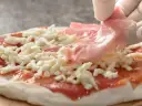 Ręka w białej rękawiczce kładzie plasterki szynki na pizzy z serem i sosem pomidorowym.